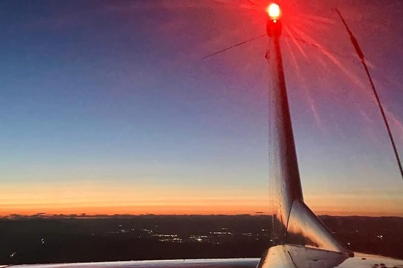 Looking back along a light aircraft’s tail fin at last light, its red anti-collision beacon lit, the wing below and town lights scattered across the ground under an orange horizon.