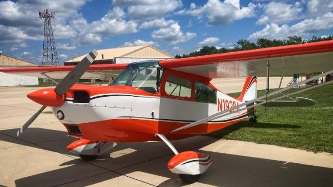 An orange-and-white Citabria 7KCAB tailwheel aeroplane parked on a concrete ramp beside a hangar row, under scattered cloud.